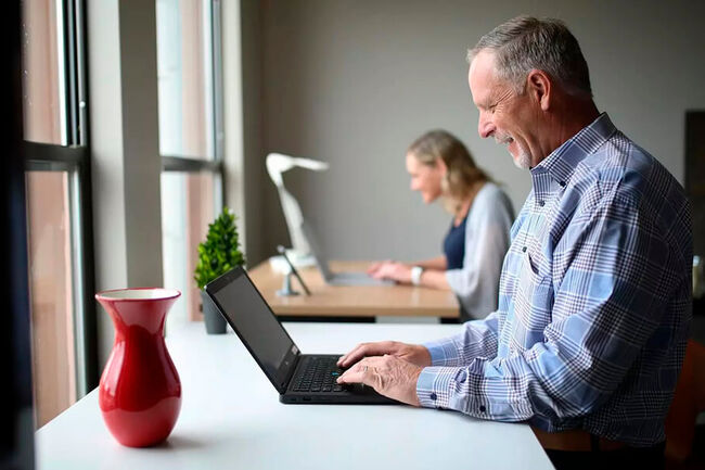 An older and a younger colleague sit next to each other, each with their own laptop